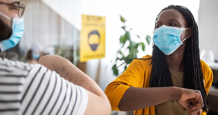 Two coworkers giving each other an elbow greeting while wearing masks in their office building.